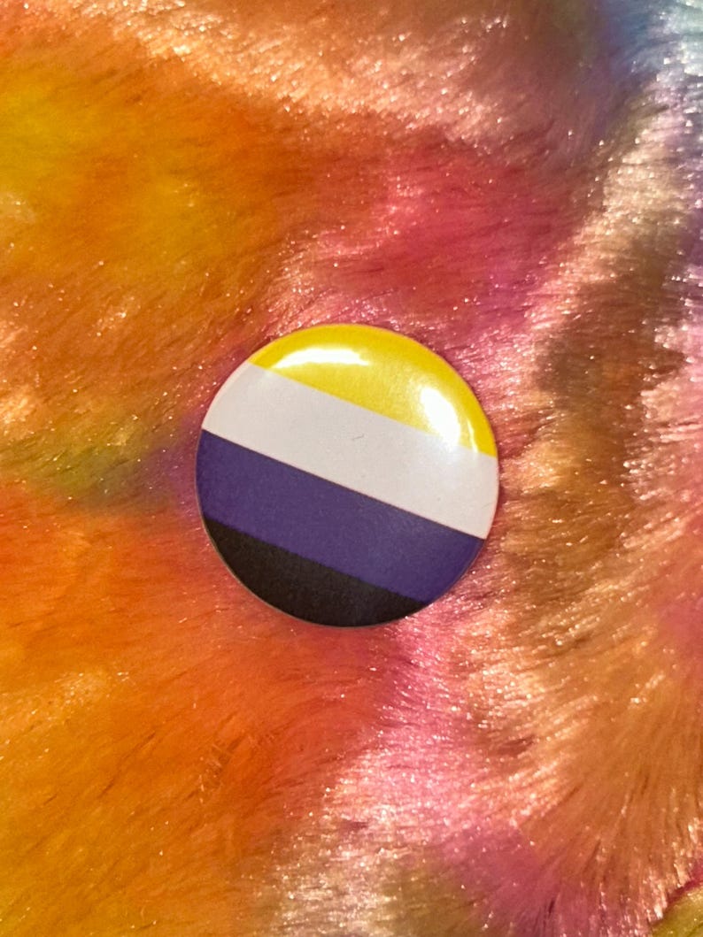 Może przedstawiać: Okrągła przypinka z flagą dumy os&oacute;b niebinarnych. Przypinka ma ż&oacute;łtą g&oacute;rę, biały pasek, fioletowy pasek i czarne dno. Przypinka jest umieszczona na kolorowym, teksturowanym tle.