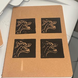 Carnet de notes artisanal en papier brun clair, orné de quatre linogravures représentant un doberman aboyant, le tout sur fond noir. #faitmain #artistique #punk #bohème #été