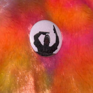 May include: A circular pinback button featuring a black and white graphic of a person wearing a hooded garment with arms raised, positioned against a vibrant, multi-colored faux fur background with shades of pink, orange, yellow, and blue.