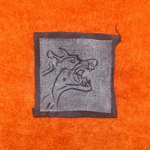 May include: A square fabric patch featuring a dark gray graphic of a snarling wolf head with an open mouth and visible teeth, set against a vibrant orange textured fabric background. The patch has a dark border and a matte finish.