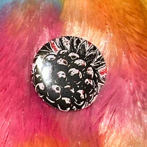 May include: A circular pin-back button featuring a high-contrast black and white linocut-style illustration of a blackberry fruit with textured leaves. The graphic design uses bold lines to define the individual drupelets of the berry.