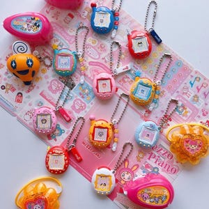 Puede incluir: Una colección de llaveros de mascotas digitales coloridos con pantallas, cadenas de metal y carcasas de plástico en varias formas y tonos, incluyendo rosa, azul, rojo, amarillo y naranja, junto a dos dijes de flores naranjas y dos estuches rosas.