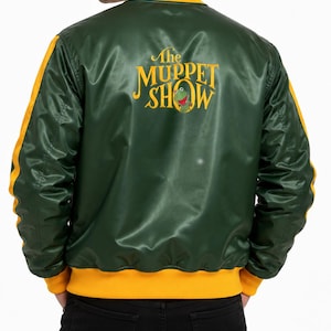 Pode incluir: Um blusão bomber de cetim verde-escuro com acabamentos canelados amarelos na gola, punhos e bainha. As costas apresentam o texto bordado a amarelo The Muppet Show, acompanhado por um pequeno gráfico do Sapo Cocas ao centro.