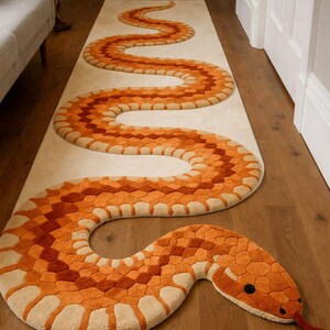 Puede incluir: Una alfombra larga con forma de serpiente naranja y beige con un patrón detallado. La alfombra tiene un aspecto texturizado y está colocada sobre un suelo de madera. La cabeza de la serpiente presenta ojos negros y una lengua roja. La alfombra es un elemento decorativo para el hogar.