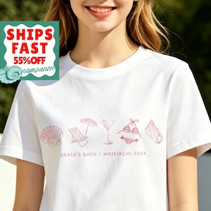 May include: White t-shirt with a pink beach-themed graphic. The design includes a seashell, beach chair, cocktail, bikini, and ice cream cone. Text on the shirt reads "GRACE'S BACH / WAIKIKI.HI 2026".
