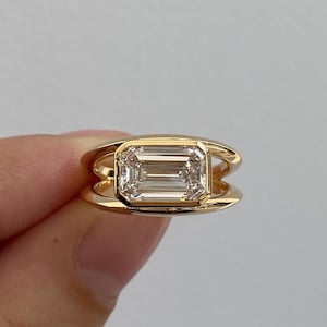 Pode incluir: Um anel de ouro com um diamante lapidação esmeralda. O diamante está engastado em uma moldura de bisel e o anel tem um design exclusivo de banda dupla. O anel é segurado, mostrando seu brilho e artesanato.