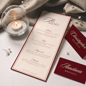 May include: A cream-colored menu card with deep red borders and text, featuring sections for starter, soup, main course, and dessert. Two matching deep red place cards display the names Christopher Rhodes and Anastasia Brooks in elegant script.