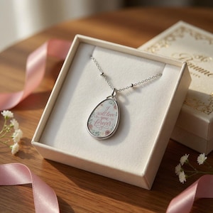 Puede incluir: Un collar plateado con un colgante en una caja de regalo blanca. El colgante tiene un diseño floral con las palabras "I will love you forever". La caja está sobre una superficie de madera con cinta rosa.