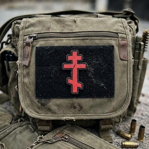 May include: An olive green canvas tactical messenger bag featuring a black rectangular patch with a red Eastern Orthodox cross. The bag has a zippered front pocket, brown leather accents, and a side compartment holding a cleaning brush tool.