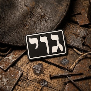 May include: A black and white patch with Hebrew text, set against a backdrop of aged leather and weathered wood. The patch features bold, stylized lettering. Rusty metal accents and a leather element add to the rustic aesthetic.