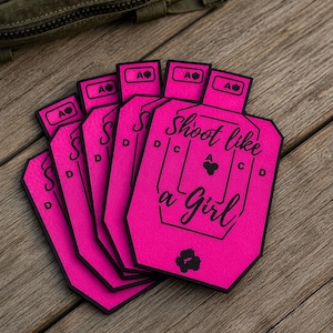 May include: A stack of bright pink paper shooting targets with black outlines and markings. Each target features the text Shoot like a Girl in black script, along with letters A, C, and D, and a small black spade symbol near the top and bottom.