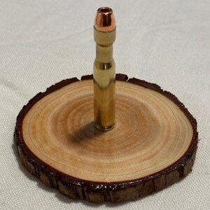 May include: A bullet-shaped object with a copper-colored tip is set in a small, round wooden base. The base is a cross-section of a tree branch, with a dark brown, bark-like edge. The wood has a natural, light brown color.