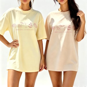 May include: Two oversized t-shirts, one pale yellow and one beige, are displayed. Each shirt features a beach-themed graphic in a reddish-pink color, along with the text "EMMA'S BACH / ST. PETE.FL 2026". The shirts are casual and suitable for a beach vacation.