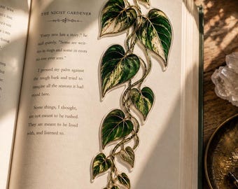 May include: A clear bookmark shaped like a trailing vine with variegated heart-shaped leaves in shades of dark green, light green, and cream. The bookmark is placed inside an open book titled The Night Gardener, page number 117 visible.