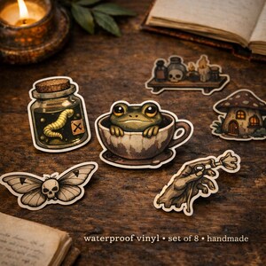 Może przedstawiać: Zestaw ośmiu wodoodpornych naklejek winylowych o fantazyjnej, gotyckiej tematyce. Zawiera żabę w filiżance, słoik z robakiem, motyla z czaszką, domek z grzybów i inne wzory. Widoczny jest tekst "waterproof vinyl • set of 8 • handmade".