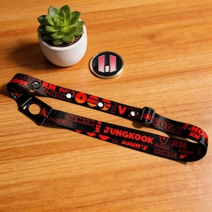 May include: Black lanyard with red text featuring names and the letters "RM", "SUGA", "J-HOPE", "JIMIN", "JUNGKOOK", and "V". The lanyard has a black buckle and a hole at the end. A small, round pin with a pink and white design is also visible.