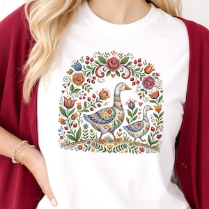 May include: White t-shirt featuring a colorful illustration of two geese surrounded by flowers and foliage. The geese have intricate patterns in blue, orange, and yellow. The design is framed by a floral archway with roses and other blooms.