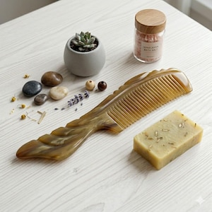 May include: A light brown, decorative comb with a carved handle, next to a bar of soap, a small potted succulent, and a jar of Himalayan bath salts. The comb is approximately 8 inches long.