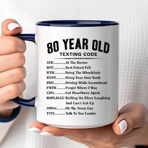 May include: White ceramic mug with a navy blue interior and handle, featuring the text "80 YEAR OLD TEXTING CODE" in black. The mug lists humorous acronyms like "ATD - At The Doctor" and "ROFLACGU - Rolling On Floor Laughing".