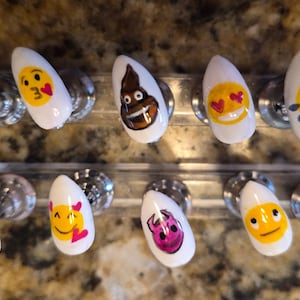 May include: A set of ten white artificial nails, each adorned with a different emoji design. The emojis include a money-eyed face, a heart-eyed face, a smiling poop, and others. The nails are displayed on a clear stand.