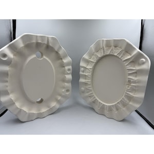 May include: Two off-white ceramic oval-shaped platters with ruffled edges. One platter has a hole and three small raised circles. The other platter has a textured design around the oval center and three small raised circles.