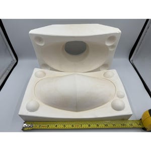 Pode incluir: Conjunto de moldes de cerâmica esbranquiçada de duas peças. A peça superior tem uma grande abertura central e três pequenas reentrâncias circulares. A peça inferior tem uma forma ovalada curva. Uma fita métrica amarela mostra cerca de 35,5 cm de comprimento.