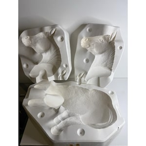 Puede incluir: Tres moldes blancos de yeso de caballos. Dos moldes muestran la cabeza del caballo y el tercer molde muestra el cuerpo del caballo. Los moldes están en un taller.