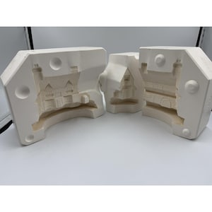 May include: Three-piece white plaster mold set for creating a miniature house. The molds are open, revealing the detailed interior and exterior features of the house, including windows, doors, and a chimney. The set is designed to be used for casting or molding.