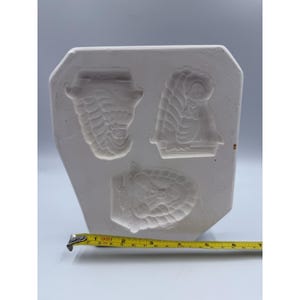 May include: A white ceramic mold with three detailed relief designs. The designs include a hat, a woman with a bonnet, and a turtle. A yellow measuring tape is placed near the mold.