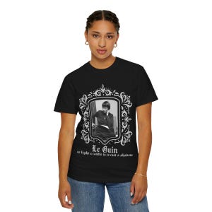 Puede incluir: Camiseta negra con un retrato en blanco y negro enmarcado en blanco. El marco tiene detalles decorativos. Debajo del retrato, el texto dice "Le Guin" y una cita.