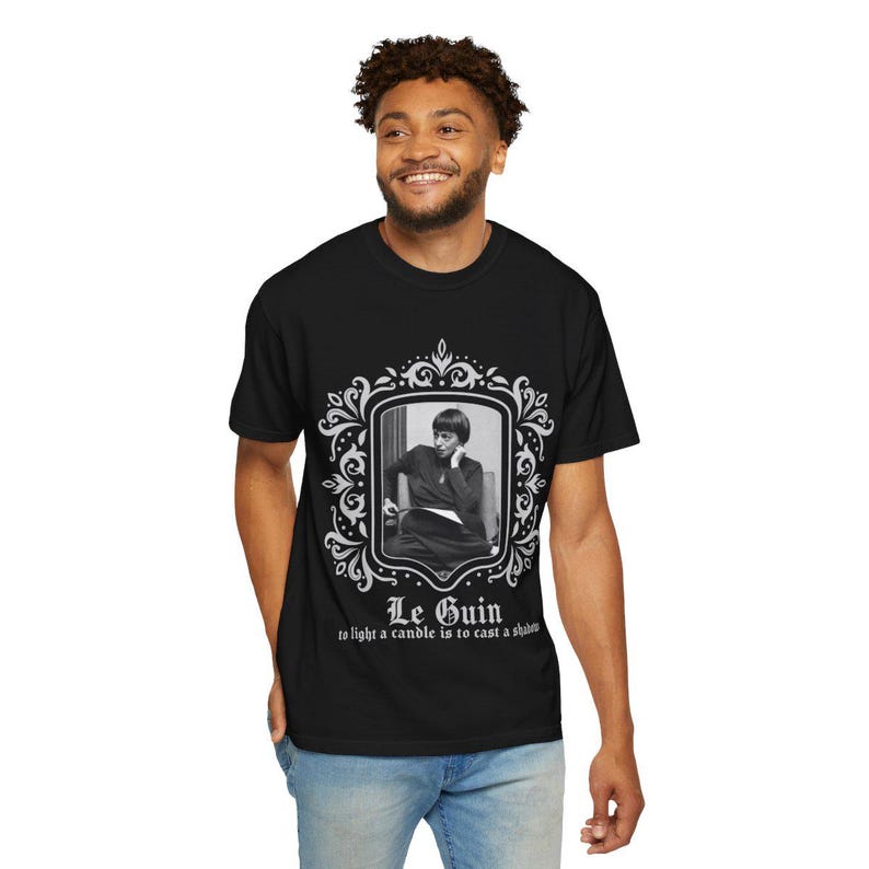 Puede incluir: Camiseta negra con un retrato en blanco y negro en un marco decorativo. El texto "Le Guin" y una cita est&aacute;n impresos debajo. La camiseta la lleva una persona.