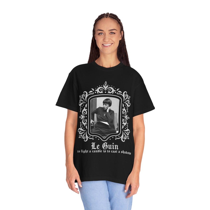 Puede incluir: Camiseta negra con un retrato enmarcado en blanco y negro de una persona. El marco es blanco con detalles ornamentados. El texto "Le Guin" y una cita est&aacute;n debajo. La camiseta est&aacute; siendo usada.