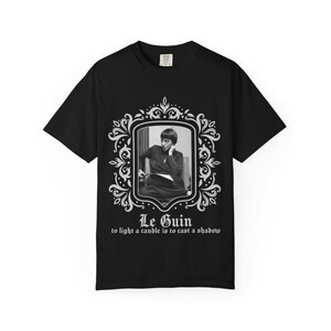 Puede incluir: Camiseta negra con un retrato en blanco y negro en un marco decorativo. El texto "Le Guin" y una cita est&aacute;n impresos debajo. La camiseta es de cuello redondo.
