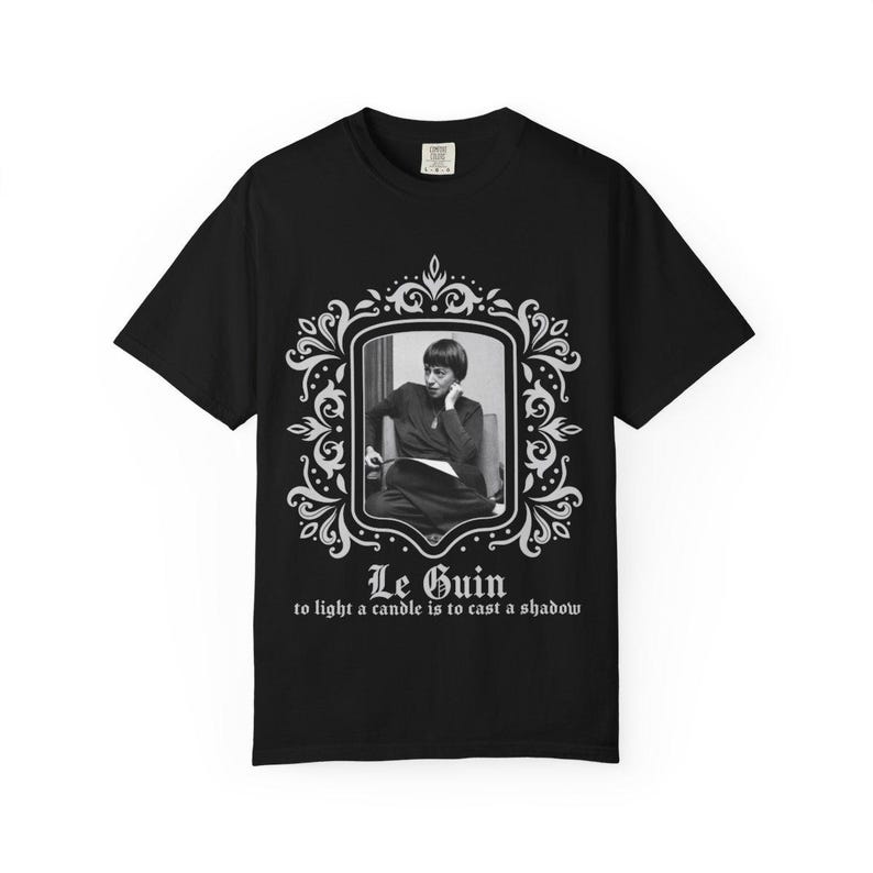 Puede incluir: Camiseta negra con un retrato en blanco y negro en un marco decorativo. El texto "Le Guin" y una cita est&aacute;n impresos debajo. La camiseta es de cuello redondo.