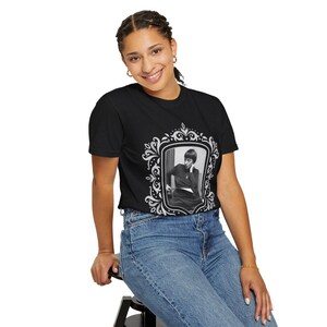 Puede incluir: Camiseta negra con un retrato enmarcado en blanco y negro. El marco tiene un dise&ntilde;o decorativo y ornamentado. La persona del retrato lleva un atuendo oscuro. La modelo lleva vaqueros azules.