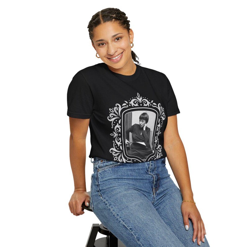 Puede incluir: Camiseta negra con un retrato enmarcado en blanco y negro. El marco tiene un dise&ntilde;o decorativo y ornamentado. La persona del retrato lleva un atuendo oscuro. La modelo lleva vaqueros azules.