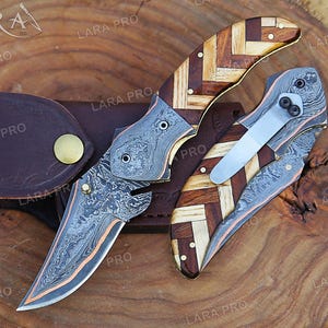 May include: Two folding knives with Damascus steel blades and handles featuring a herringbone pattern of light and dark wood. One knife is open, showing the patterned metal, while the other is closed with a silver pocket clip attached to the side.