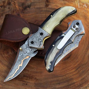 May include: Two folding knives with Damascus steel blades featuring a wavy copper inlay. The handles are crafted from polished horn with brass pins. One knife is open, revealing the patterned metal, while the other is closed, showing a silver pocket clip.