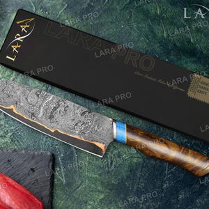 May include: A handmade Damascus knife with a patterned blade and a wooden handle. The knife has a blue and gold accent near the handle. The knife is displayed with its black box, which has the text "LARA PRO".