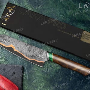 May include: A Damascus knife with a patterned blade and a wooden handle. The blade has an orange accent and a green section near the handle. The knife is displayed with its black box, which has gold lettering and the words "HANDMADE DAMASCUS KNIFE".