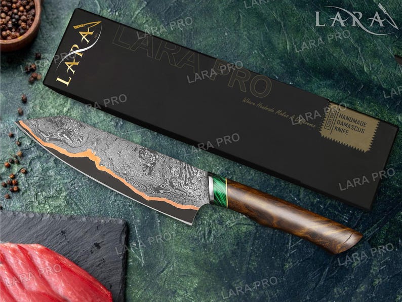 May include: A Damascus knife with a patterned blade and a wooden handle. The blade has an orange accent and a green section near the handle. The knife is displayed with its black box, which has gold lettering and the words "HANDMADE DAMASCUS KNIFE".