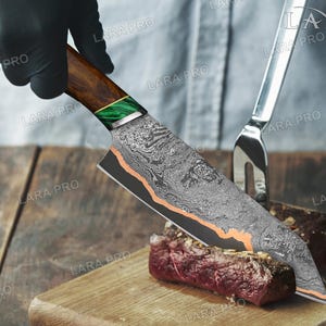 May include: A Damascus steel chef's knife with a wooden handle and green accents is cutting a piece of raw meat on a wooden cutting board. A silver fork is in the background. The knife has a patterned blade and copper-colored accents.