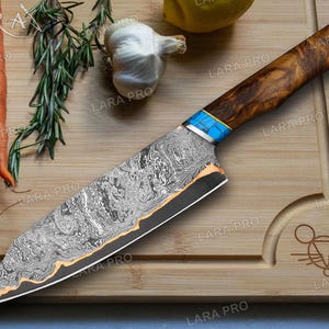 May include: A Damascus steel chef's knife with a wooden handle and blue accents. The blade has a unique pattern and a gold-colored edge. The knife rests on a wooden cutting board with carrots, rosemary, and garlic.