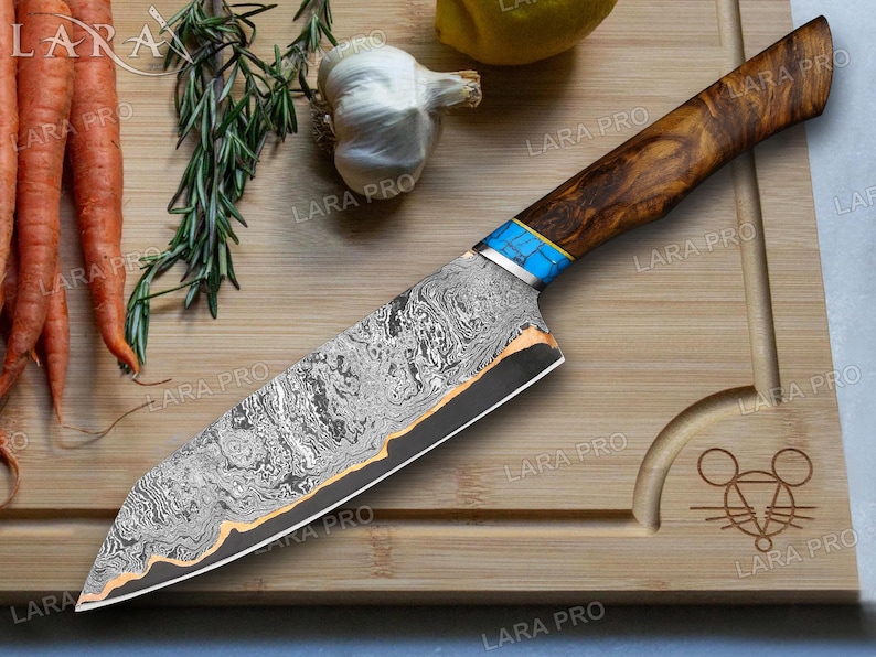 May include: A Damascus steel chef's knife with a wooden handle and blue accents. The blade has a unique pattern and a gold-colored edge. The knife rests on a wooden cutting board with carrots, rosemary, and garlic.