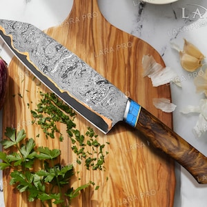 May include: A Damascus steel chef's knife with a patterned blade and a wooden handle with blue accents. The knife rests on a wooden cutting board with chopped herbs and eggplants.