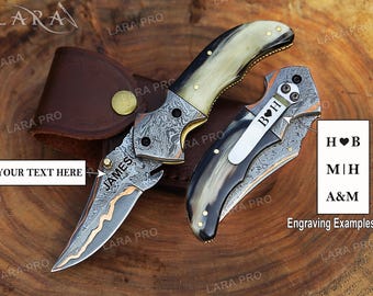 May include: Two folding knives with Damascus steel blades and horn handles. One blade features the engraved text "JAMES". A metal pocket clip on the second knife is engraved with "B♥H". A brown leather sheath with a gold snap button is visible.