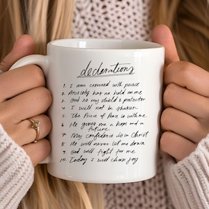 May include: White ceramic mug with the word "declarations" in script. The mug features a list of ten affirmations in black ink. The mug is held by a person wearing a cream-colored sweater.