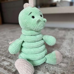 May include: A light green crocheted plush toy shaped like a dinosaur with a segmented body, small arms, and light beige accents on the feet, back, and head. The toy features black safety eyes and a soft, textured yarn construction throughout its form.