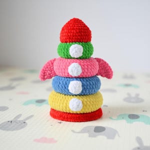 May include: A handmade crochet stacking toy in a rocket ship design. The toy features a red base and top, with yellow, blue, pink, and green rings. Each ring has a white circular accent. The toy is designed for babies and toddlers.