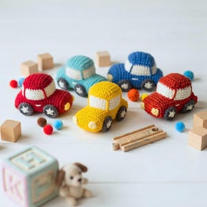 May include: A collection of five small, handmade crochet toy cars in red, yellow, light blue, and dark blue. Each car features white windows and wheels with star-shaped centers. Scattered wooden blocks, small felt pom-poms, and a wooden track piece.